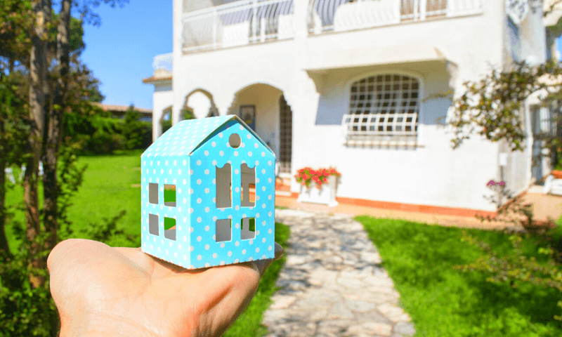 Una mano che tiene una piccola casa in miniatura davanti a una grande casa bianca
